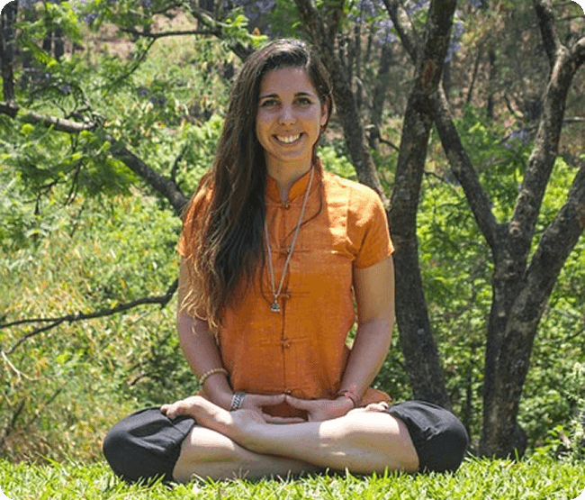 Woman in orange clothing sitting in meditation pose outdoors in a forest setting